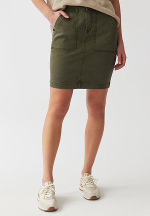 Olive green denim skirt with two front pockets, fitted design, and a hemline reaching mid-thigh, paired with beige sneakers.
