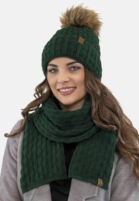 Sötétzöld kötött sapka műszőrme pom-pommal, egy hozzá illő, textúrált sállal kombinálva. Bordás mintázattal és egy kis márkás címkével rendelkezik.