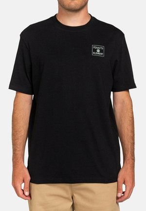 Man wearing a black short-sleeve T-shirt with a small white "Peanuts Element" logo on the left chest and beige pants.