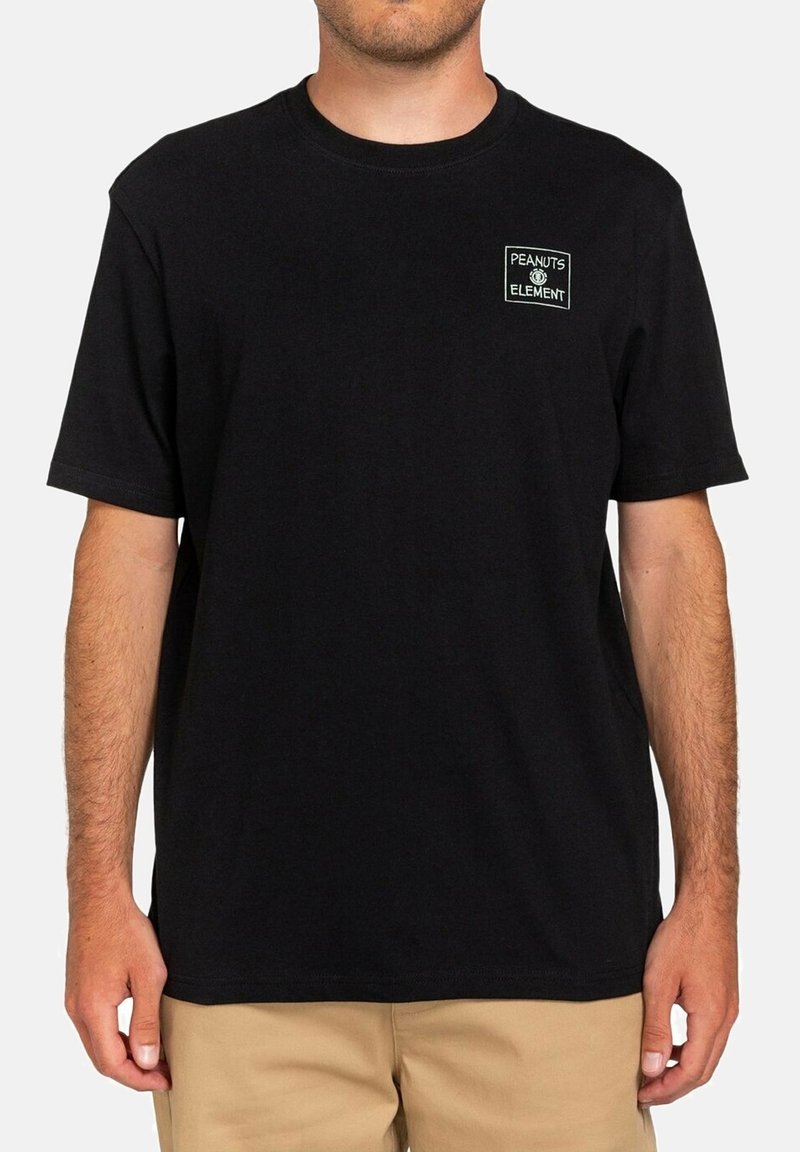 Homme portant un T-shirt noir à manches courtes avec un petit logo blanc « Peanuts Element » sur le côté gauche de la poitrine et un pantalon beige.