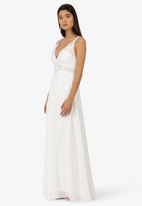 Weißes Kleid mit tiefem V-Ausschnitt, Spitzenoberteil und Spaghettiträgern, das in einen bodenlangen, leichten Rock übergeht.