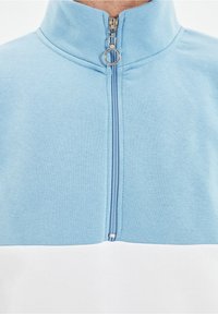 Detailní záběr na světle modrou a bílou mikinu na zip se stříbrným kruhovým zipem u výstřihu.