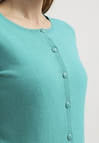 Turkos kofta gjord av stickat material, med rund halsringning och knappstängning framtill med matchande knappar längs placket.