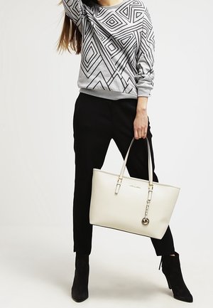 Femme portant un pull gris à motifs géométriques et un pantalon noir, tenant un grand sac cabas blanc Michael Kors avec un porte-clés logo.