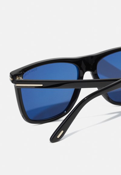 Occhiali da sole in plastica nera con lenti azzurre. Presentano un design rettangolare elegante e dettagli dorati sulla montatura.