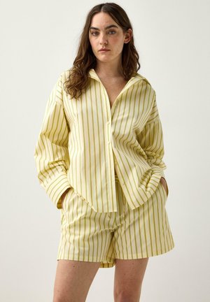 Donna con capelli lunghi castani che indossa una camicia a righe verticali gialle e bianche con bottoni e pantaloncini coordinati, mani nelle tasche, sfondo neutro.
