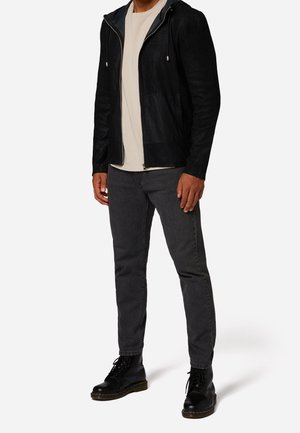 Homme portant un sweat à capuche noir zippé, un t-shirt beige, un jean gris foncé et des bottes noires à lacets, debout devant un fond blanc uni.