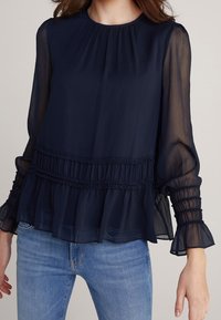 Marinefarbene Bluse mit langen Ärmeln aus transparentem Stoff, mit Falten-Detail, gerafftem Bund und Puffärmeln, kombiniert mit hellblauen Jeans.