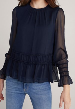 Marinefarbene Bluse mit langen Ärmeln aus transparentem Stoff, mit Falten-Detail, gerafftem Bund und Puffärmeln, kombiniert mit hellblauen Jeans.