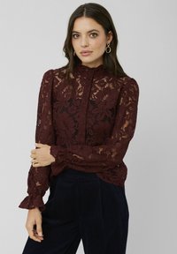 Blouse en dentelle bourgogne avec un col à volants haut, des manches longues et une fermeture à boutons. Motif floral transparent avec des poignets évasés et des détails délicats.