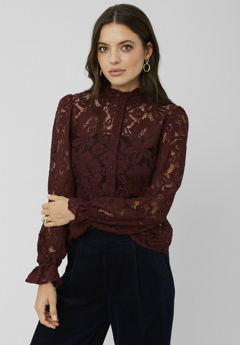 Blouse en dentelle bourgogne avec un col à volants haut, des manches longues et une fermeture à boutons. Motif floral transparent avec des poignets évasés et des détails délicats.