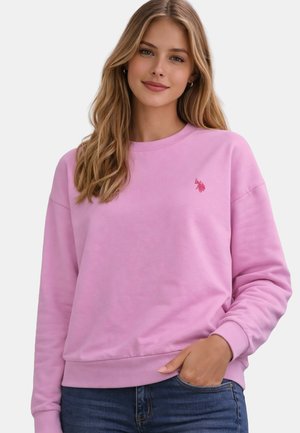 Young woman with long blonde hair wears a light purple sweatshirt with small embroidered logo, paired with blue jeans, hand in pocket.