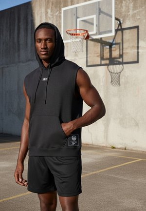 Atletisk mann i svart ermeløs hettejakke og shorts står på utendørs basketballbane med kurv og betongvegg bak seg.