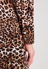 Šaty s leopardím potiskem, které mají hnědou a černou barevnou paletu, dlouhé rukávy, zapínání na knoflíky na manžetách a hladkou texturu látky.