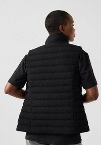 Gilet imbottito nero con colletto alto, caratterizzato da una superficie testurizzata e un design trapuntato orizzontale. Indossato sopra una maglietta nera a maniche corte.