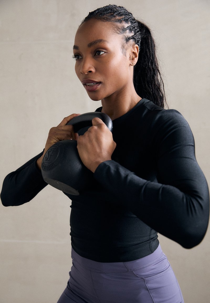 Kvinde med fletninger, der løfter en kettlebell tæt på sit bryst, iført en sort langærmet trøje og lilla leggins, indendørs.