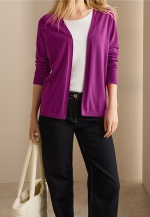 Femme portant un cardigan violet ouvert, un haut blanc, un jean noir, tenant un sac fourre-tout blanc matelassé sur un fond beige.