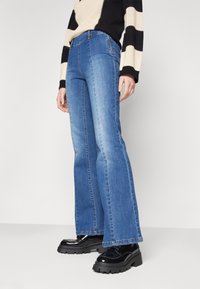 Ausgestellte blaue Denim-Jeans mit mittlerem Waschgang, hoher Taille, geraden Beinen und genähten Details entlang der Nähte.