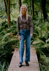 Blůza s leopardím potiskem, světle modré džíny s vysokým pasem, černé kotníkové boty; modelka stojí na dřevěné chodníku obklopená zelení.