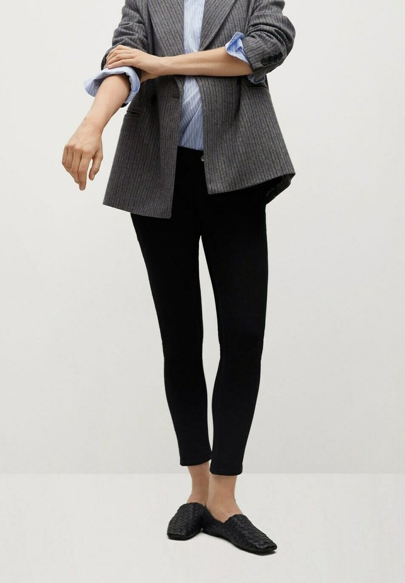 Personne portant un blazer gris à fines rayures, une chemise bleu clair retroussée, un pantalon noir ajusté et des chaussures noires tissées sans lacets, debout sur un sol blanc uni.