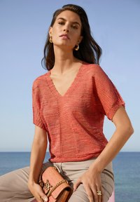 Koralfarbener Strickpullover mit V-Ausschnitt und kurzen Ärmeln, kombiniert mit hellbeigen Hosen und einer rosa geflochtenen Handtasche mit goldener Kette.