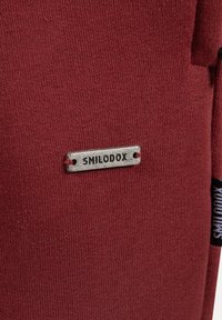 Czerwona dzianina z małą prostokątną metalową metką wygrawerowaną napisem "SMILODOX", przymocowaną czerwonym przeszyciem.
