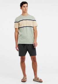 Chemise à manches courtes en vert clair et beige avec une bande marine sur la poitrine ; associée à un short sombre et des sandales marron.
