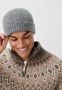 Gorro de canalé gris sobre una persona sonriente que lleva un suéter de lana con cremallera y un patrón de rombos en tonos beige, marrón y crema.