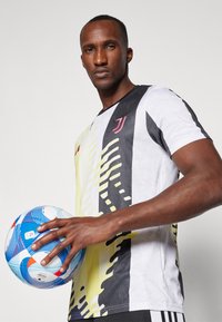 Maillot à manches courtes blanc et jaune à motifs avec des accents noirs. Le joueur tient un ballon de soccer bleu et blanc, mettant en avant une prise détaillée.