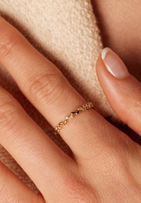 Gouden ring met een op bladeren geïnspireerd ontwerp, met kleine kristallen ingelegd, gedragen aan een vinger, tegen een textuur-rijke, lichte achtergrond.