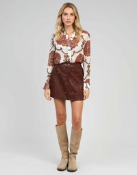 Femme portant un chemisier à manches longues à motifs, une mini-jupe en cuir marron cloutée et des bottes en suède beige jusqu'aux genoux, se tenant contre un fond uni.