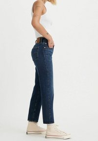 Jeans de mezclilla azul de talle alto y pierna recta con un diseño clásico de cinco bolsillos, remaches de cobre y un parche de cuero marrón en la cinturilla.