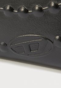 Čierny kožený výrobok s textúrovaným povrchom, zaoblenými nýtmi a embosovaným logom. Hladké prevedenie s jemným leskom a detailným vzorom.