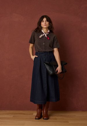 Femme portant un haut marron à manches courtes, un pantalon large foncé, des bottes marron, une écharpe en soie, tenant un sac à main noir tissé, debout sur un sol en bois.