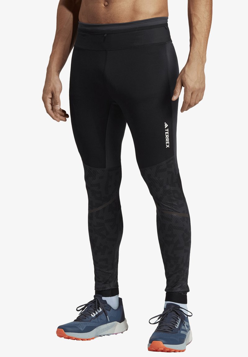 Black stretchy athletic leggings with textured panels, featuring a logo on the thigh and a fitted design. Worn with blue and orange running shoes.