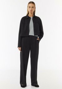 Zwarte cropped jas met knoopsluiting, twee voorzakken, gecombineerd met een gestreept shirt en hoge zwarte broeken. Glad stoffen textuur.