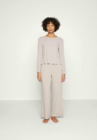 Femme portant un haut ample à manches longues beige clair et un pantalon évasé, debout pieds nus, sur un fond uni clair.