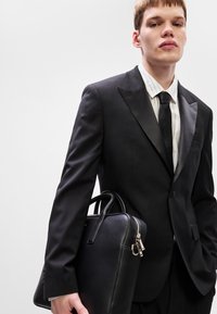 Μαύρο κοστούμι με σατέν επικάλυψη, άσπρη ριγέ πουκαμίσα και μαύρη γραβάτα. Κρατάει ένα δομημένο μαύρο δερμάτινο χαρτοφύλακα με ασημένιο φερμουάρ.