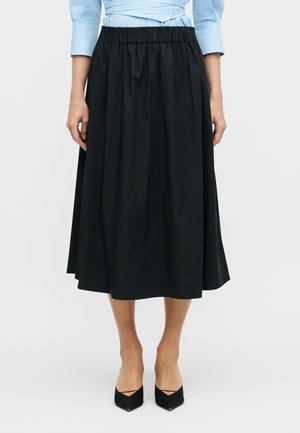 Žena v čiernej midi sukni s elastickým pásom, v kombinácii s čiernymi lodičkami s ostrou špičkou a svetlomodrým topom.