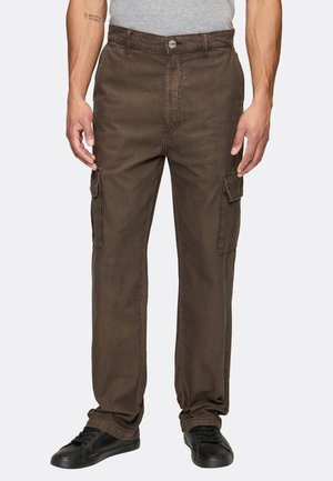 Mand iført brune cargobukser med klaplommer på siderne, grå skjorte og sorte sneakers, der står mod en ensfarvet baggrund.