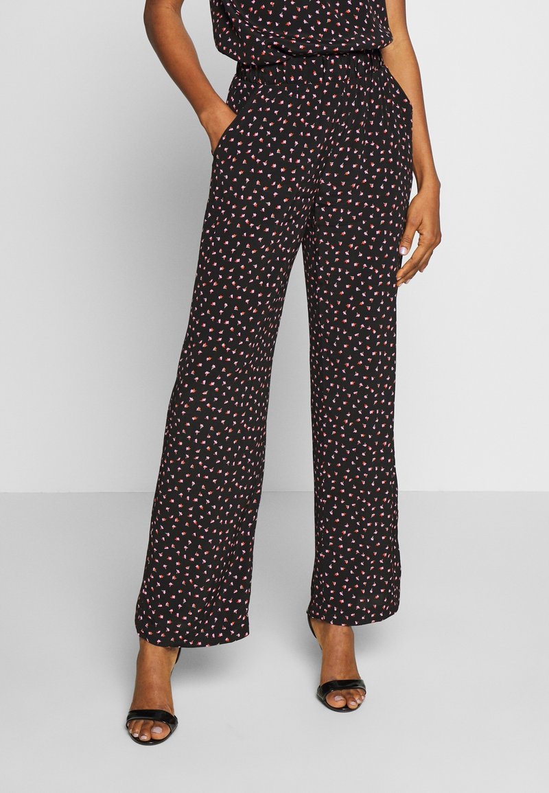 Femme portant un pantalon noir à jambes larges avec un petit imprimé floral rouge et blanc, des talons ouverts noirs, debout avec une main dans la poche.