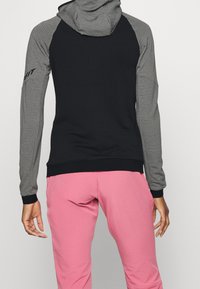 Felpa sportiva nera e grigia con cappuccio, texture a costine sulle maniche, polsini neri e banda inferiore, abbinata a pantaloni sportivi rosa.
