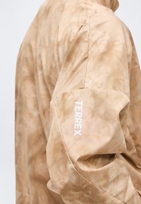 Beež värviga tie-dye jopevarrukas, mille ülaosas on valge "TERREX" logo vertikaalselt trükitud.