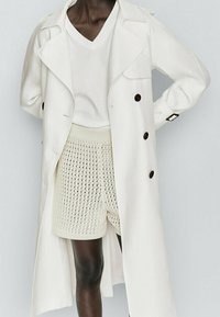 White trench coat with large lapels and double-breasted buttons, layered over a white V-neck t-shirt and textured cream shorts.