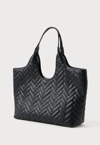 Fekete bőr bevásárlótáska steppelt halszálka mintával és két hurok fogantyúval, egy sima fehér háttér előtt állva.