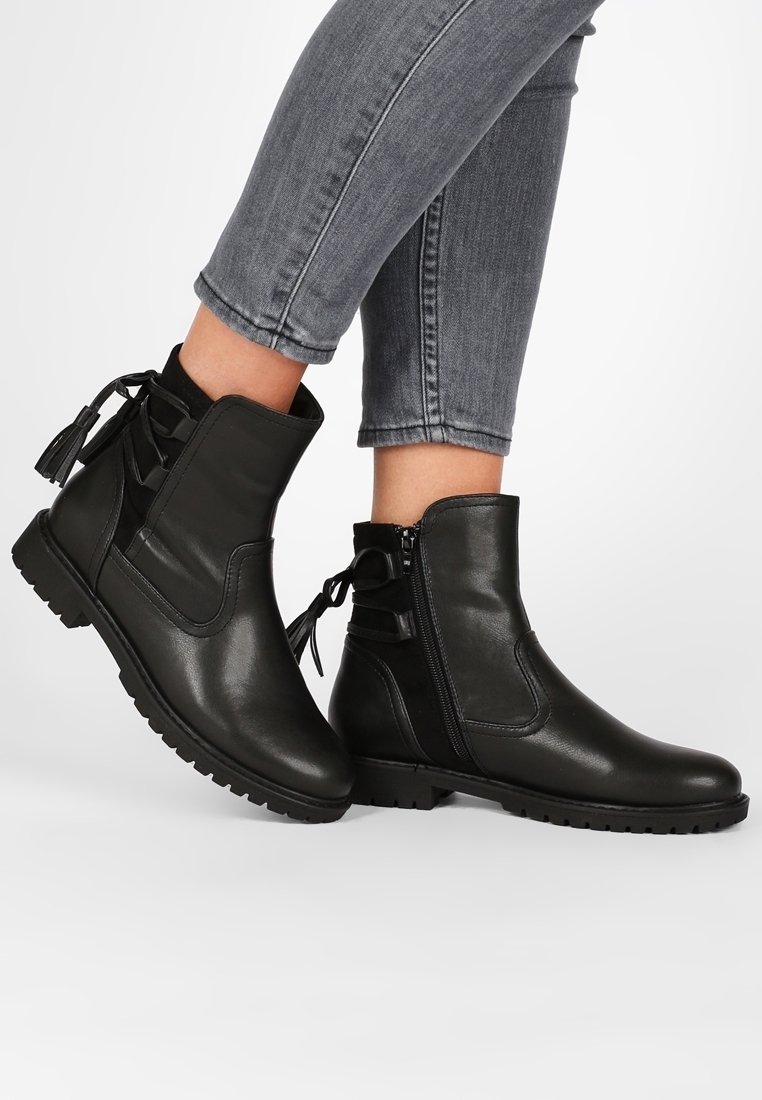 Bottes en cuir noir hauteur cheville avec fermetures éclair latérales, pampilles décoratives à l'arrière, portées avec un jean gris ajusté.