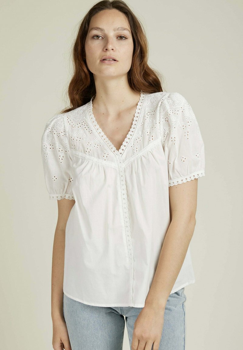 Blouse en coton blanc avec des détails en broderie à jours sur les manches bouffantes, un décolleté en forme de V et une bordure en dentelle festonnée. Associée à un jean bleu clair.