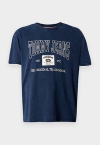 Tamsiai mėlynas medvilninis marškinėliai trumpomis rankovėmis, apvalia iškirpte ir baltais užrašais: "TOMMY JEANS," "1985," "ORIGINALUS PREKĖS ŽENKLAS," ir etikete.