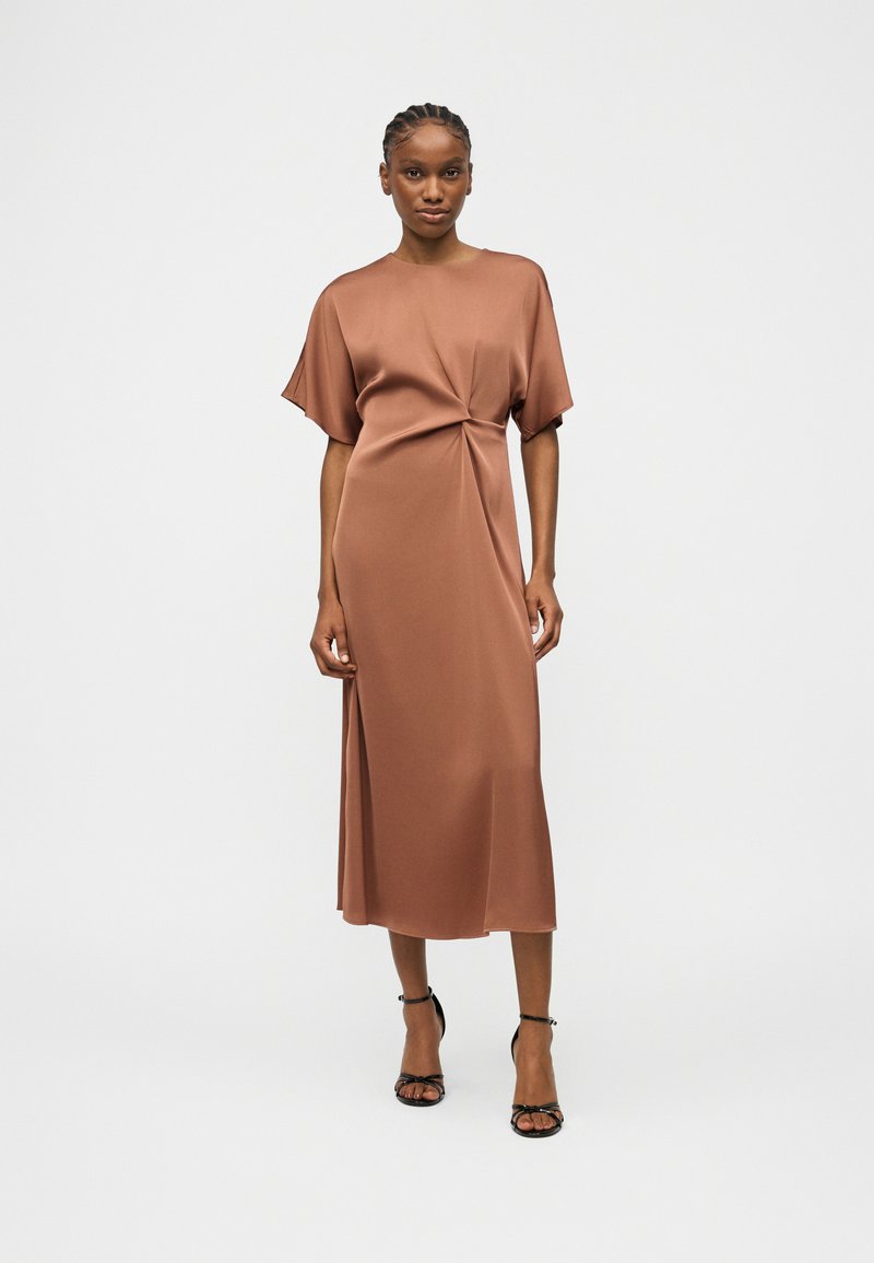 Γυναίκα που φοράει καφέ σατέν μίντι φόρεμα με κοντά μανίκια, πλευρική σχισμή και λεπτομέρεια κόμπου στη μέση, και μαύρες ψηλοτάκουνες γόβες με λουράκι στον αστράγαλο.