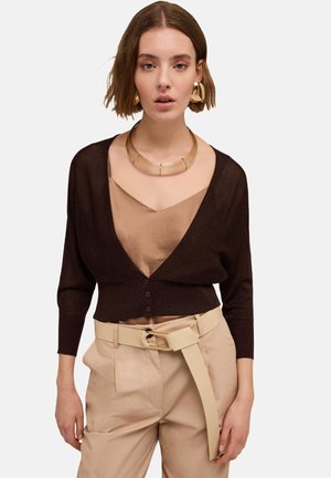 Cardigan marron court avec un décolleté en V profond, fermeture à boutons et manches longues. Porté sur un top en satin beige et associé à un pantalon beige.